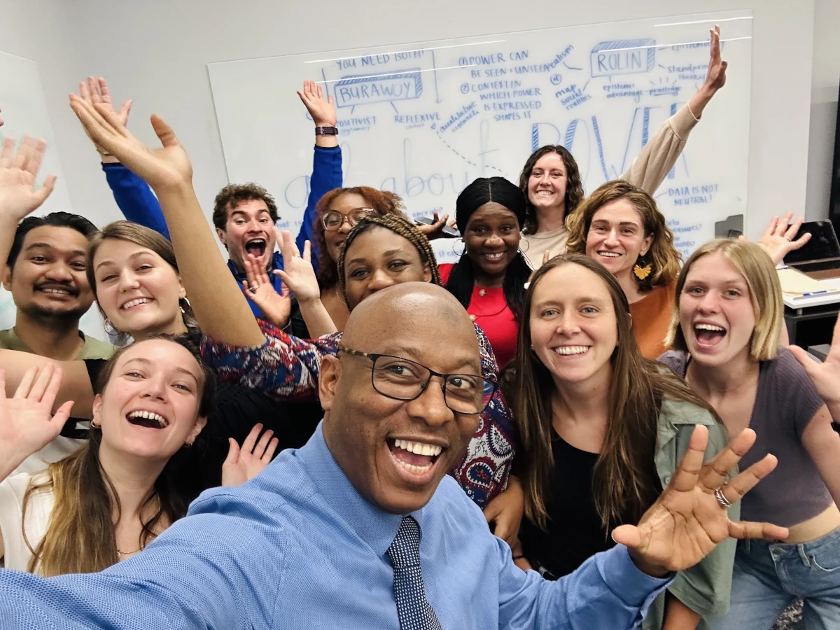 Smiling students and a faculty member taking a group selfie in a classroom, raising their hands and posing enthusiastically.